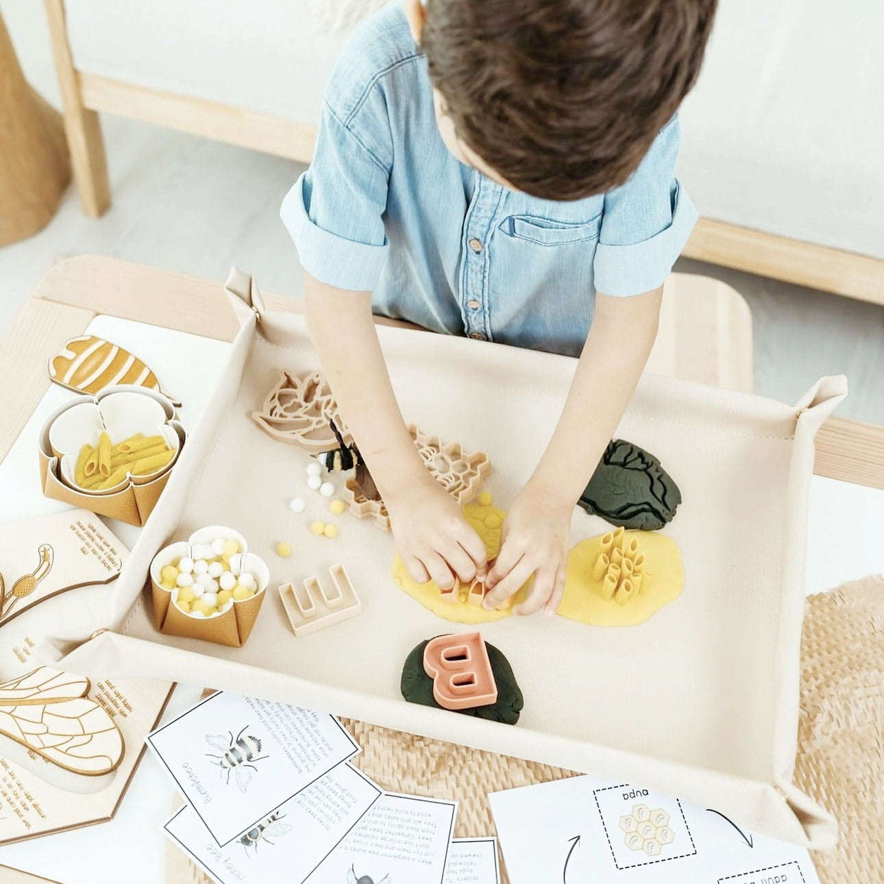 Bandeja sensorial de piel vegana color arena con niño manipulando plastilina, letras de madera y elementos de abejas sobre una mesa infantil.
