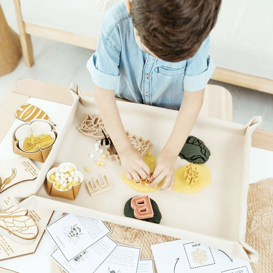 Bandeja sensorial de piel vegana color arena con niño manipulando plastilina, letras de madera y elementos de abejas sobre una mesa infantil.